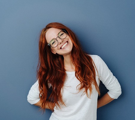 A young woman with red hair and glasses tilting her head to the side and smiling after finishing her treatment with Invisalign in Bowie